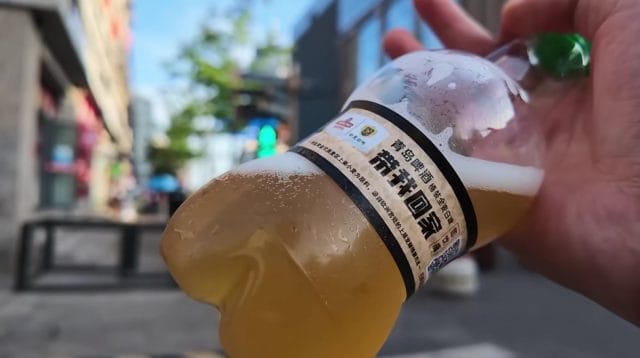 Tangan memegang botol plastik berisi bir berwarna keemasan dengan label aksara Mandarin
