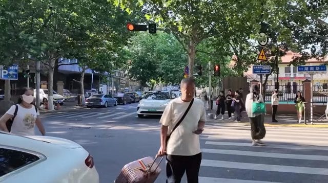 Seorang turis pria membawa koper besar menyeberangi jalan raya dengan aman di kota Qingdao
