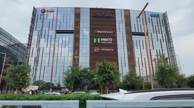 Fasad megah gedung perkantoran kaca bertingkat tinggi di kawasan pusat bisnis Gurugram yang menampilkan papan nama berbagai merek perusahaan multinasional terkemuka