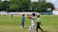Sekelompok anak bermain kriket di tanah lapang berumput hijau.