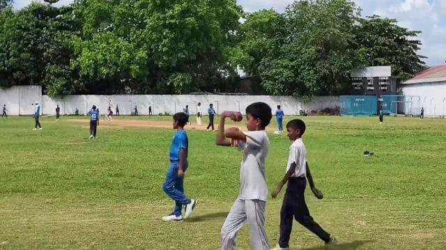 Sekelompok anak bermain kriket di tanah lapang berumput hijau.