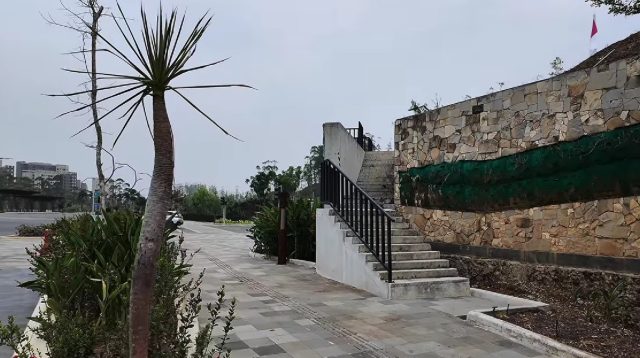 Tangga beton eksterior berpegangan besi di area fasilitas publik dengan dinding batu alam estetis.