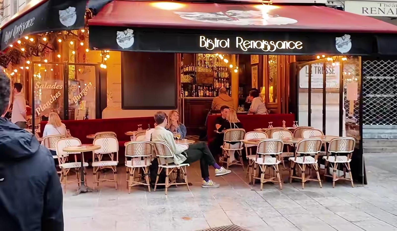 Suasana luar ruangan sebuah kafe kecil di Paris bernama Bistrot Renaissance dengan beberapa pelanggan duduk di kursi rotan.