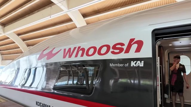 Kepala Kereta Cepat Whoosh berlogo merah terparkir di peron stasiun dengan petugas berdiri di pintu masuk.