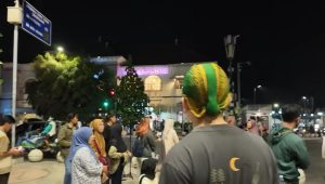 Seorang pria tampak dari belakang mengenakan blangkon hijau bercorak emas khas gaya Yogyakarta di keramaian kota.