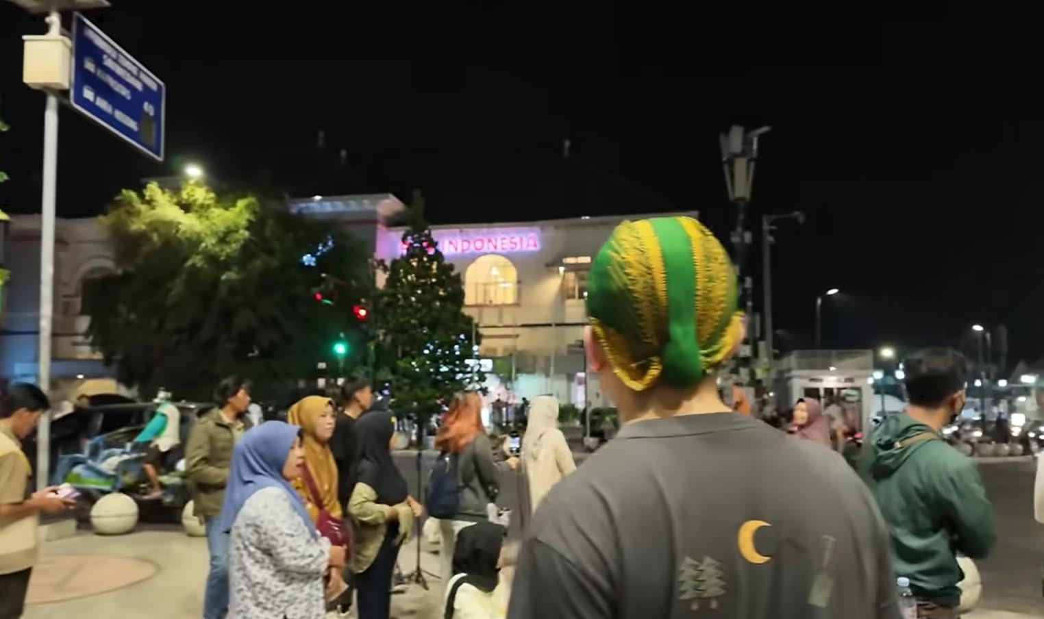 Seorang pria tampak dari belakang mengenakan blangkon hijau bercorak emas khas gaya Yogyakarta di keramaian kota.