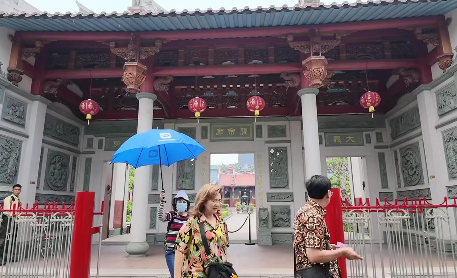 Pengunjung memegang payung biru berdiri di depan gerbang Vihara Satya Budhi Bandung yang megah.